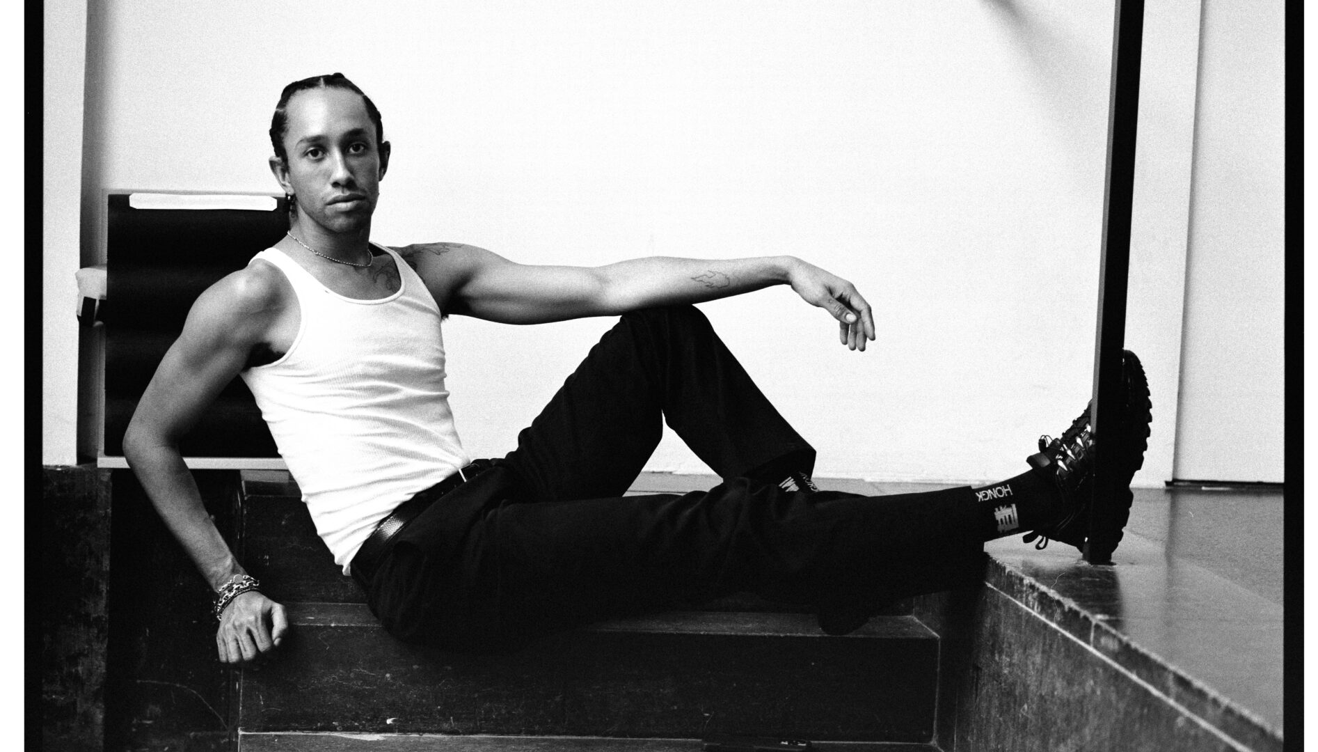 Black and white photo of a Black man in a white tank top and black pants, sitting on a stair.