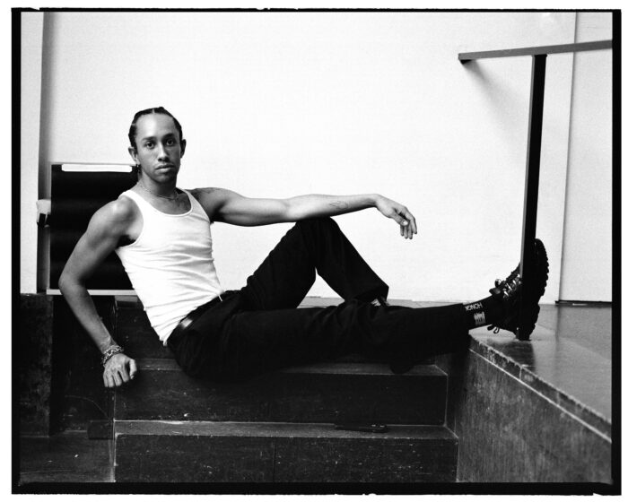 Black and white photo of a Black man in a white tank top and black pants, sitting on a stair.