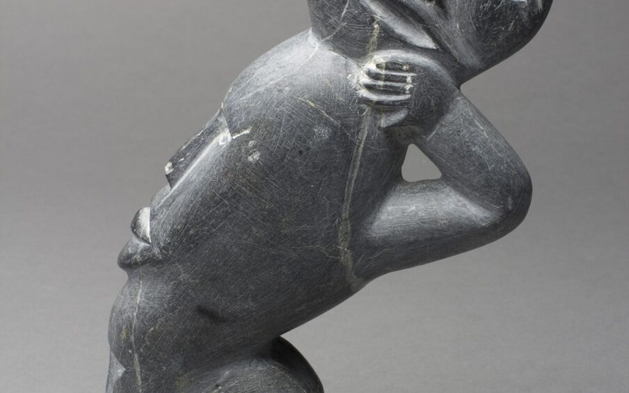 Image Description: Spirits. Silas Aittauq. 1986. Serpentine carving 9 in x 4 1/2 in x 1 3/4 in. Sleek gray carving of a spirit with multiple faces. From the angle of the photograph - the body is facing to the right, but the head is turned to face the camera. The face is round with almond shaped eyes and a thin nose carved into it. A slightly opened mouth lies behind a raised strip under the nose. It has bangs that are parted in the middle and angle to each side of the head. The body is leaning forward, right arm bent at the elbow with the hand holding the chin of the turned face. The legs are bent at the knees and there are rounded feet below. The back and space behind the legs has the profile of two additional faces. Long thin noses and plump lips are visible. There are blemishes in the material and scratches across the sculpture. A prominent light colored scratch runs from the right eye, over the lips and chin, over the finger, and down to the armpit.