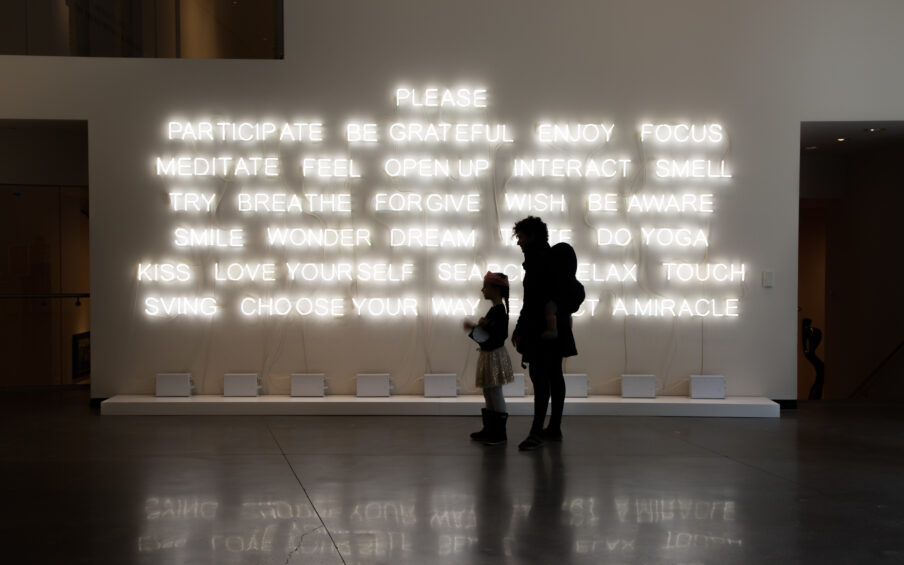 Photo of the silhouette of an adult and a child against a neon word art installation.