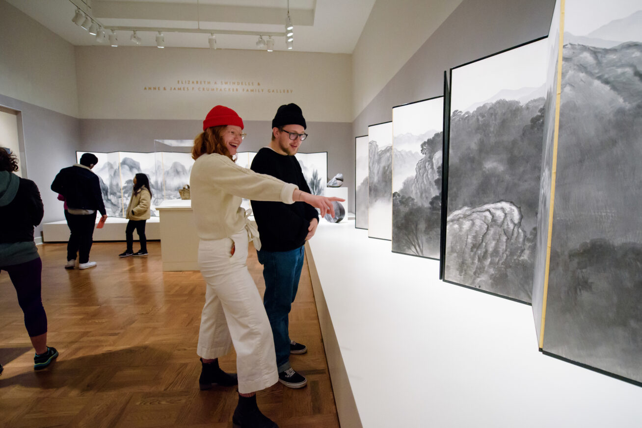 Two smiling people wearing glasses and beanies in a gallery. Woman with curly red hair pointing at an artwork.
