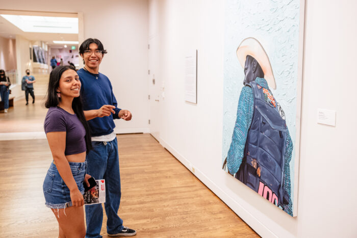 Two people in an art gallery in front of a painting looking toward the camera.