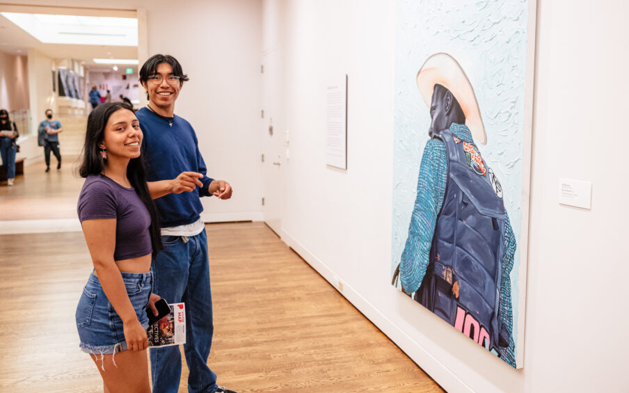 Two people in an art gallery in front of a painting looking toward the camera.