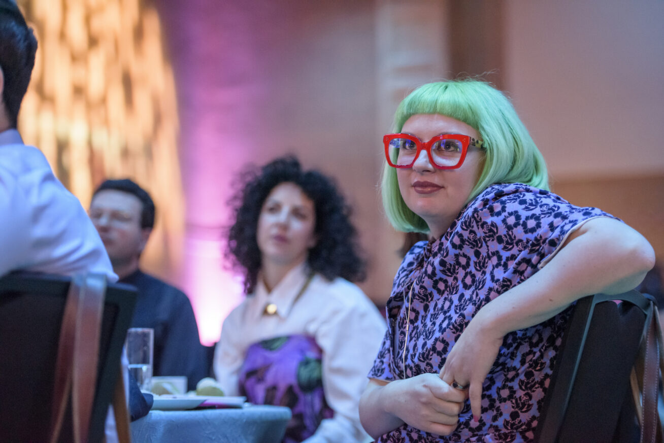 Photo of woman in the foreground with green hair and pink glasses.