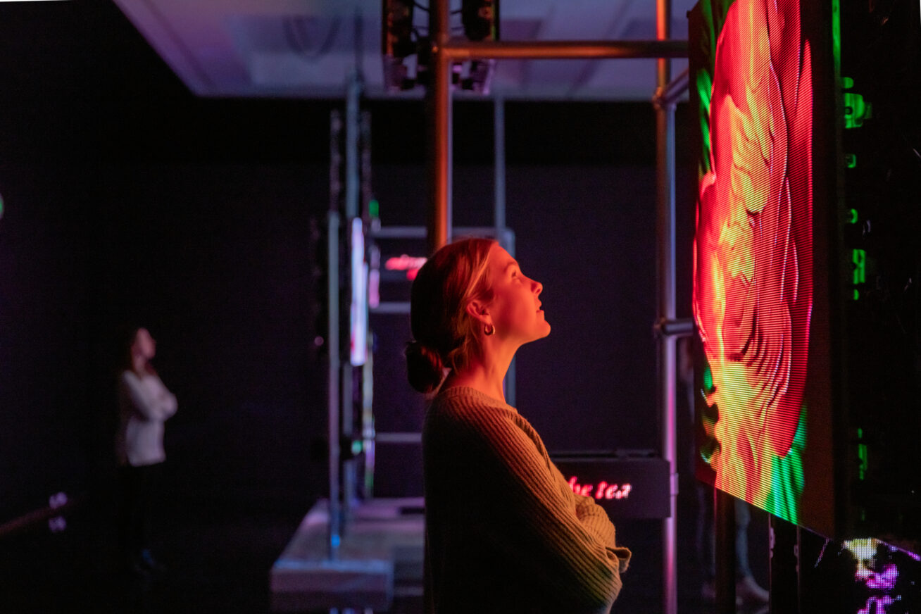 A woman with blond hair in a bun, looking up a video screen showing an AI version of a flower