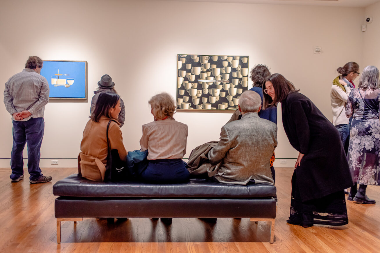 A group of people in a museum gallery, some are sitting on a bench and they are in conversation.