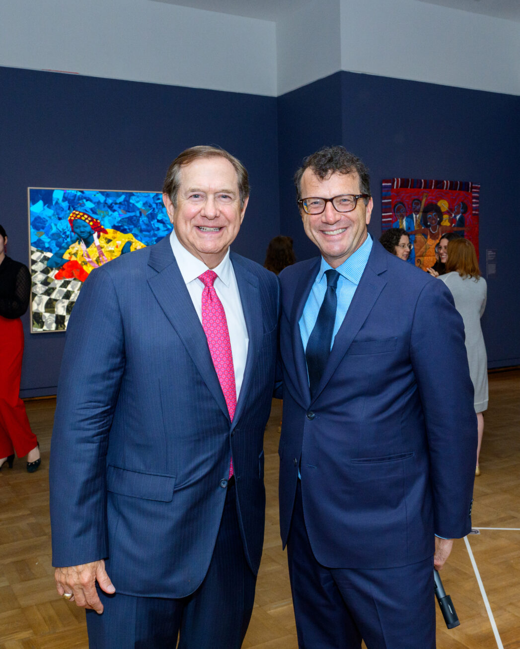 Two smiling white men in blue suit coats, standing in an art gallery.