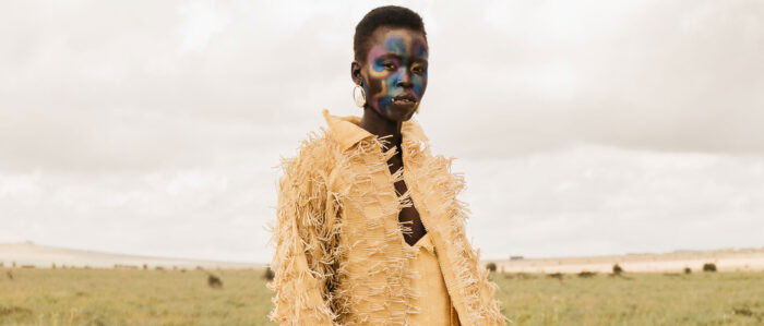 Photo of a black model with iridescent face paint and a yellow fringe jacket standing against a cloudy sky and grassy landscape