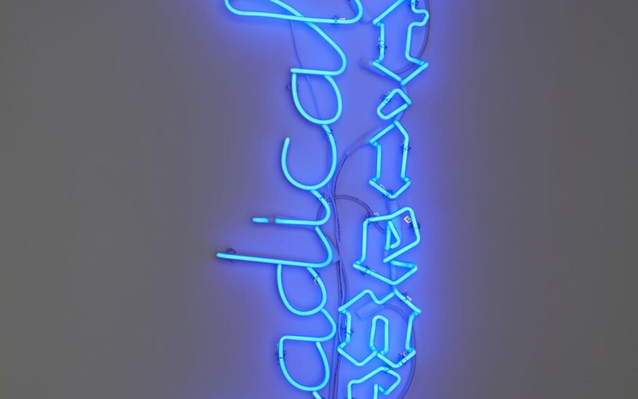 Blue neon sign with the word “Radical” in cursive positioned to the left of “Patience” in Gothic black letter and adjacent to a white analog clock that displayed 12:10 at the time of the photo.