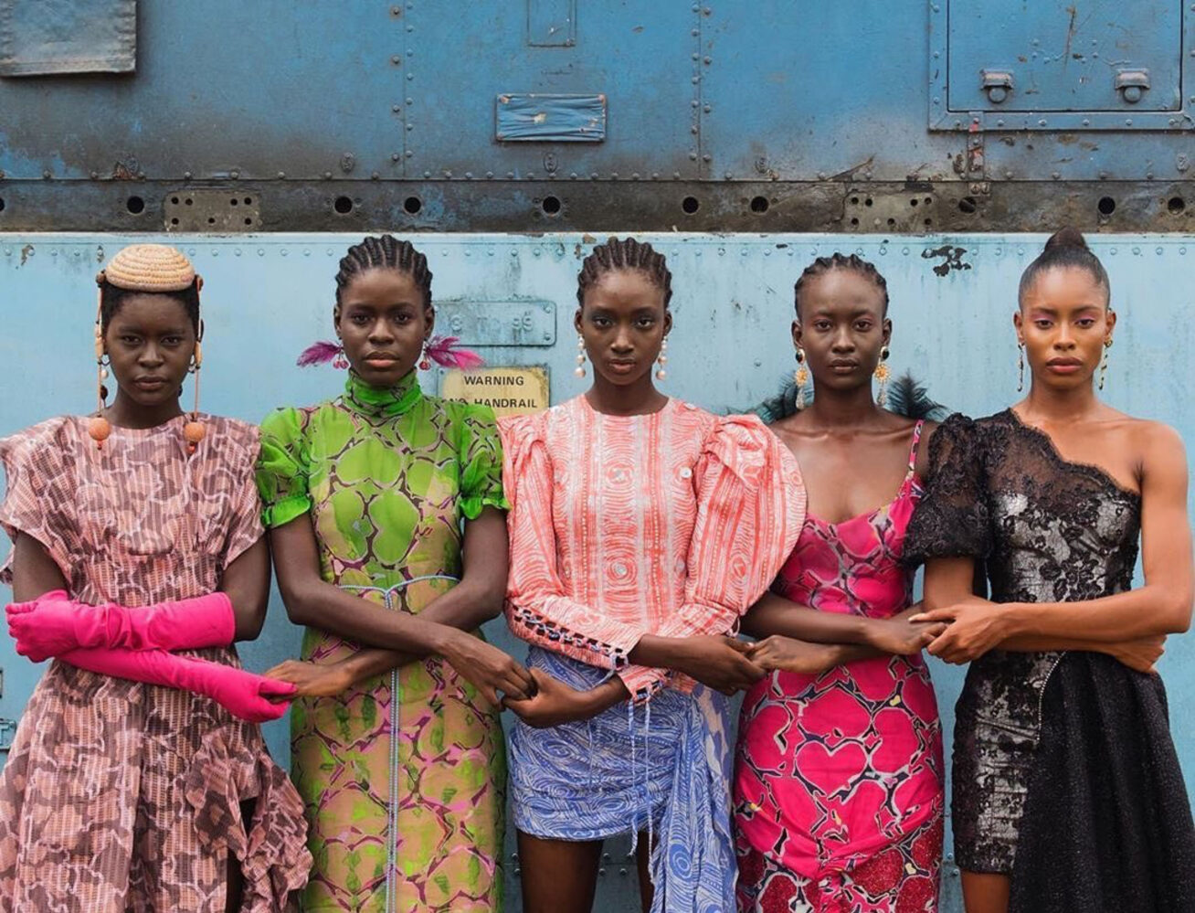 Five black female models with arms crossed and holding hands against a blue metallic wall