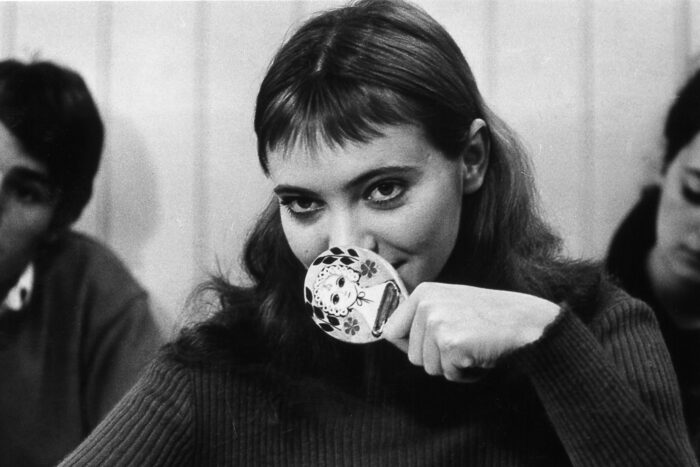 A black and white film still of Anna Karina holding a small mirror up in front of her mouth.