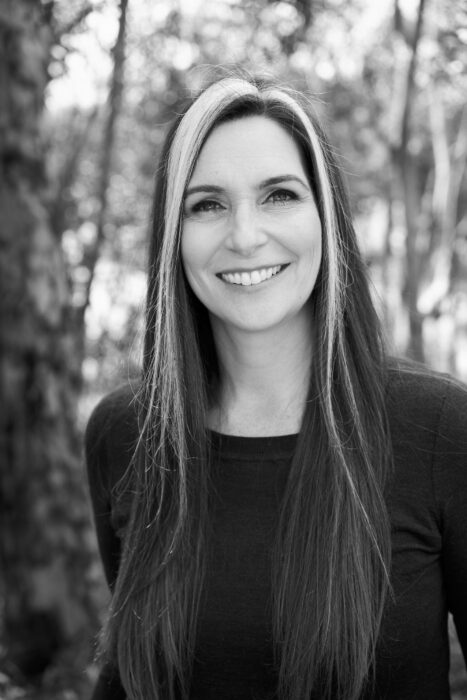 Black and white portrait of a smiling woman with long, straight dark hair with white streaks in front in front of trees.