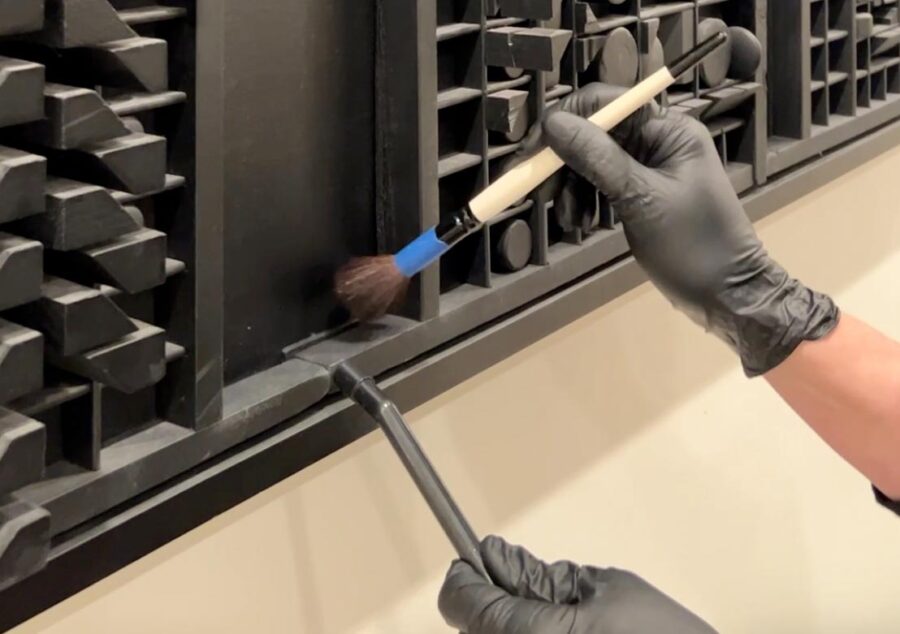 Two hands in black latex gloves brushing and vacuuming a black wooden artwork by Louise Nevelson