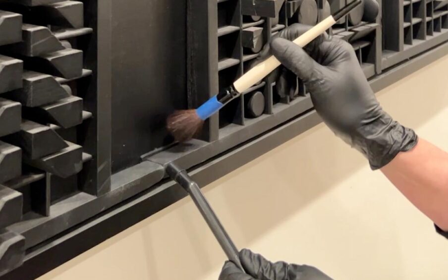 Two hands in black latex gloves brushing and vacuuming a black wooden artwork by Louise Nevelson.
