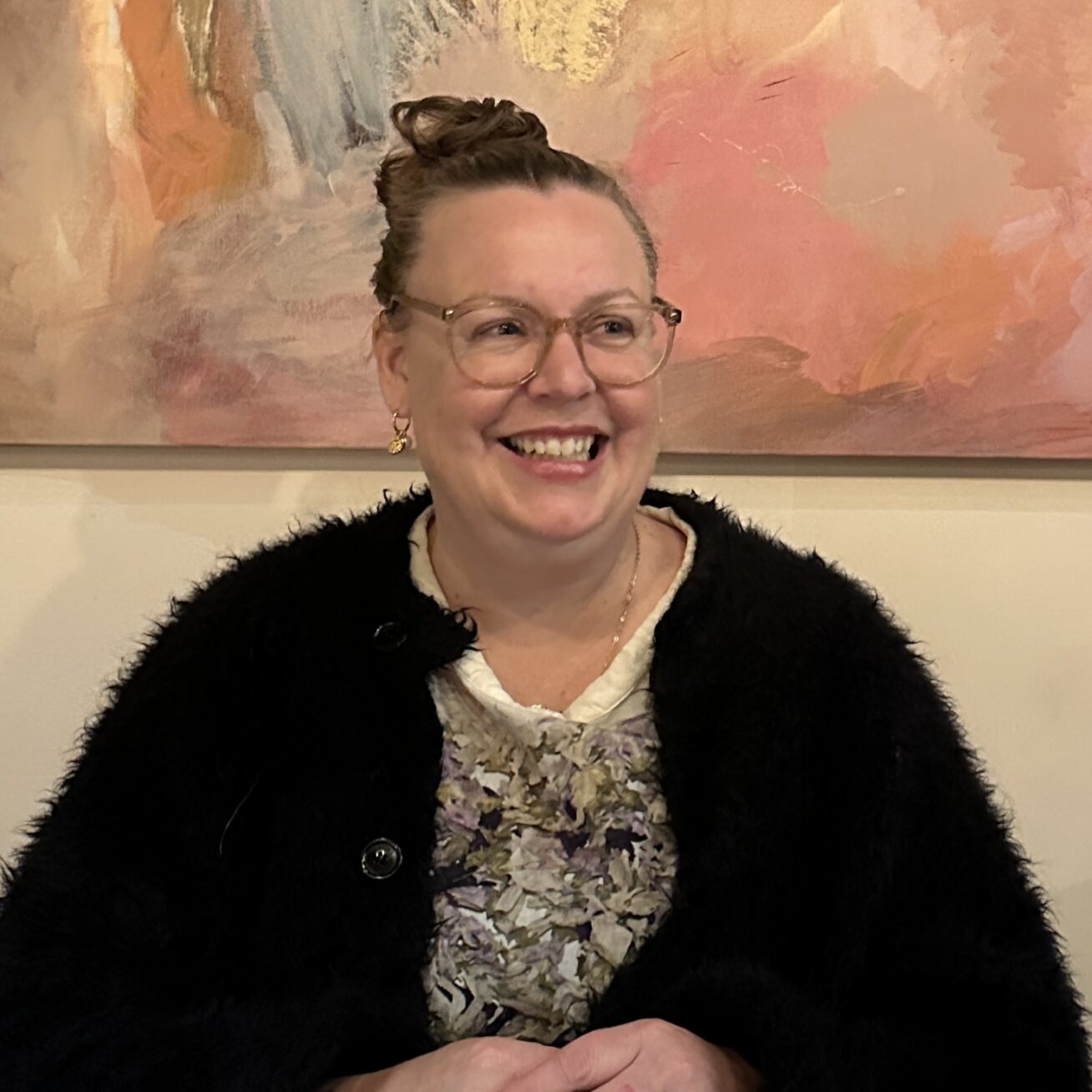 Picture of a smiling woman with her hair pulled up in a bun, wearing eyeglasses, sitting in front of a painting.