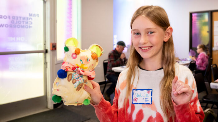 A girl with long blond hair, wearing a white and red sweater, smiling and holding up a handcrafted creature.
