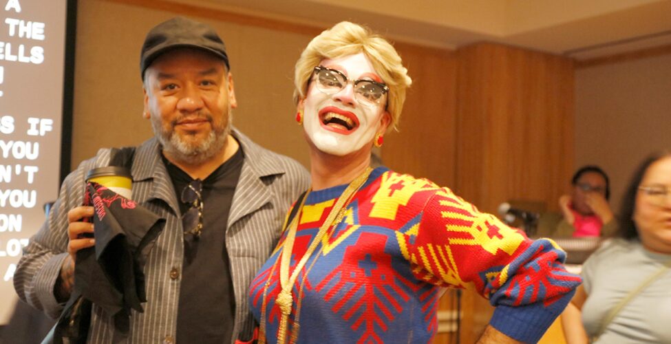 Man in a black hat with a grey beard, black t-shirt and grey and white striped coat standing next to a drag queen with a blond wig, white face paint, and a blue, red, and yellow sweater.