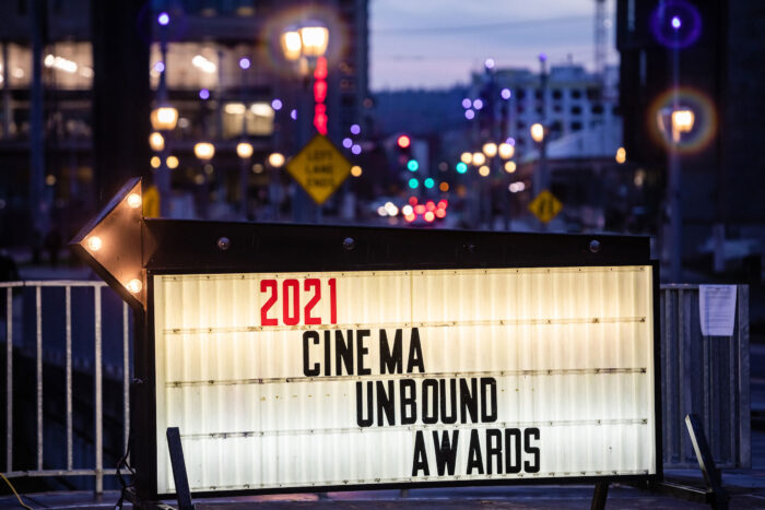 Lit up sign that says "2021 Cinema Unbound Awards" at dusk with cityscape and lights behind it