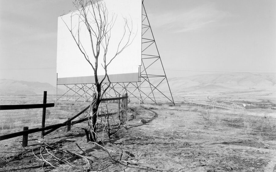 Image description: The Dalles, Oregon, Dan Powell, 1989, gelatin silver print, image: 12 11/16 x 15 7/8 inches; sheet: 16 x 19 7/8 inches. A black and white photograph featuring a drive-in movie screen and a bare tree amid an austere hilly, landscape. The movie screen occupies the upper left quadrant of the photo and shows the screen in a three-quarter view. A triangular structure supports the screen and its crossbar construction creates further triangular shapes. A small leafless tree stands near the screen and its darker branches cover most of the surface of the white screen. A dark wooden post and wire screen fence starts at center left and juts into the space behind the tree. The surrounding ground is strewn with bare branches and scrub grass. In the distance at right center and even with the bottom of the movie screen, the Dalles Bridge crosses a portion of the Columbia River. Low-slung, far-off hills bisect the photo in half. The top half of the photo shows a pale sky with light, wispy clouds.
