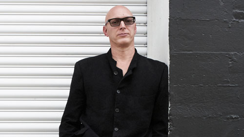 Photo of a white man in a black suit and sunglasses leaning against a black wall with a metal door behind him.