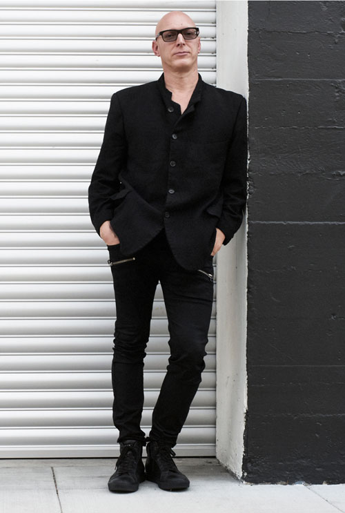 Photo of a white man in a black suit and sunglasses leaning against a black wall with a metal door behind him.