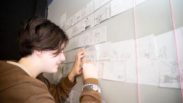 A kid wearing a brown sweatshirt drawing on paper pinned up on a whiteboard.