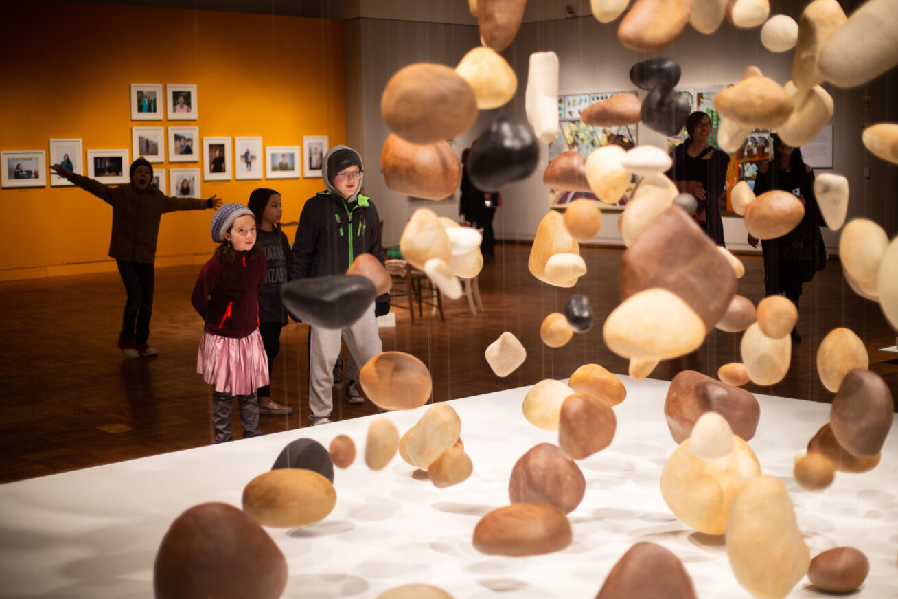Children in winter clothing standing in a gallery and looking at an art installation of rocks hanging from the ceiling