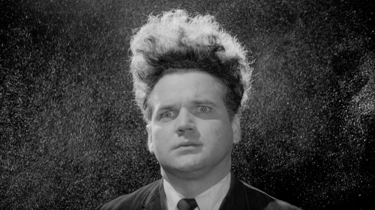 Black and white film still of a man with hair sticking straight up with a worried look on his face.