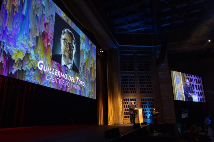 Guillermo del Toro speaking at podium with projections behind and to the side of him.