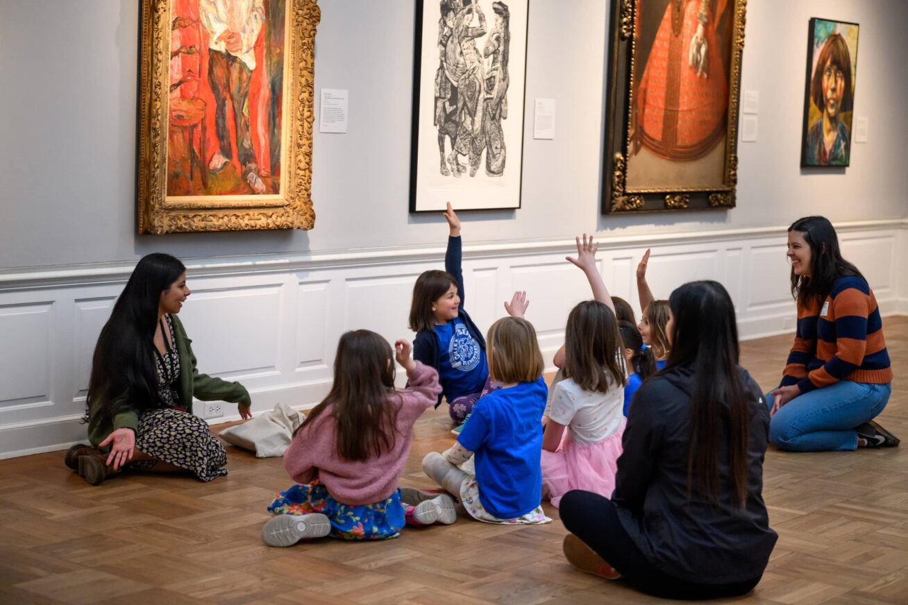 A group of children sitting on a gallery floor with hands raised to ask questions.