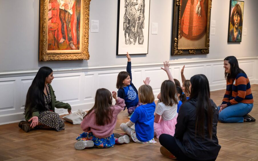A group of children sitting on a gallery floor with hands raised to ask questions.