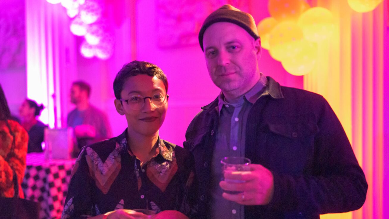 Two people holding drinks in front of a bright pink and yellow backdrop.