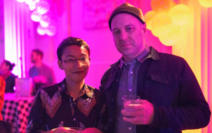 Two people holding drinks in front of a bright pink and yellow backdrop.
