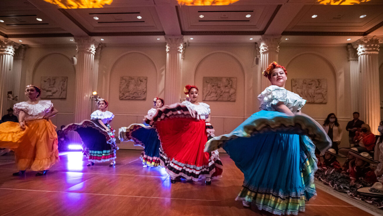 Five dancing young women in brightly colored traditional Mexican Jalisco dresses with flowers in their hair.
