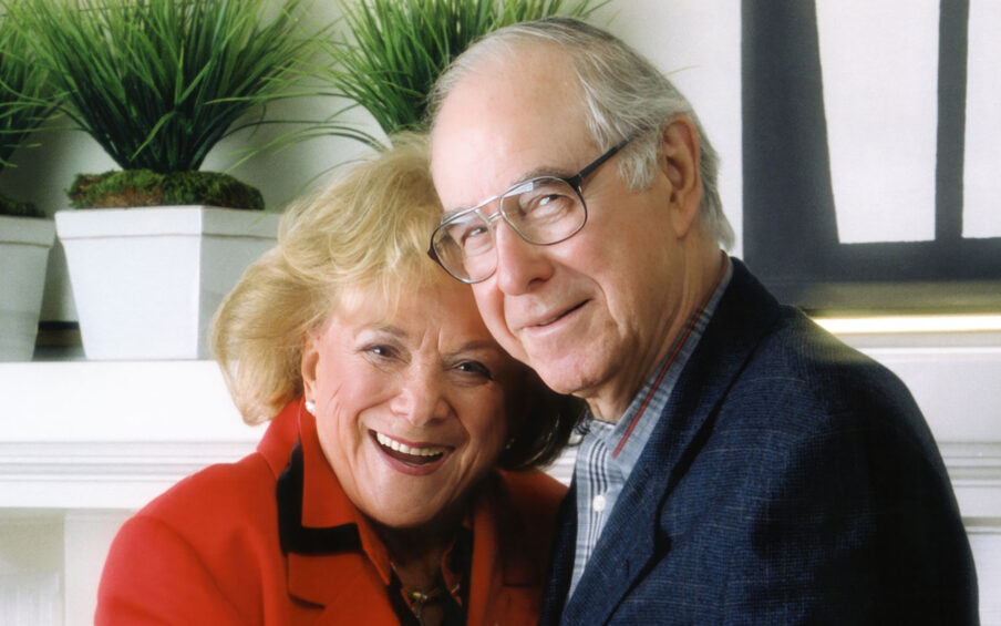 A smiling older white woman with blond hair, wearing a red jacket, standing next to a smiling older white man with eyeglasses and a blue suit coat on.