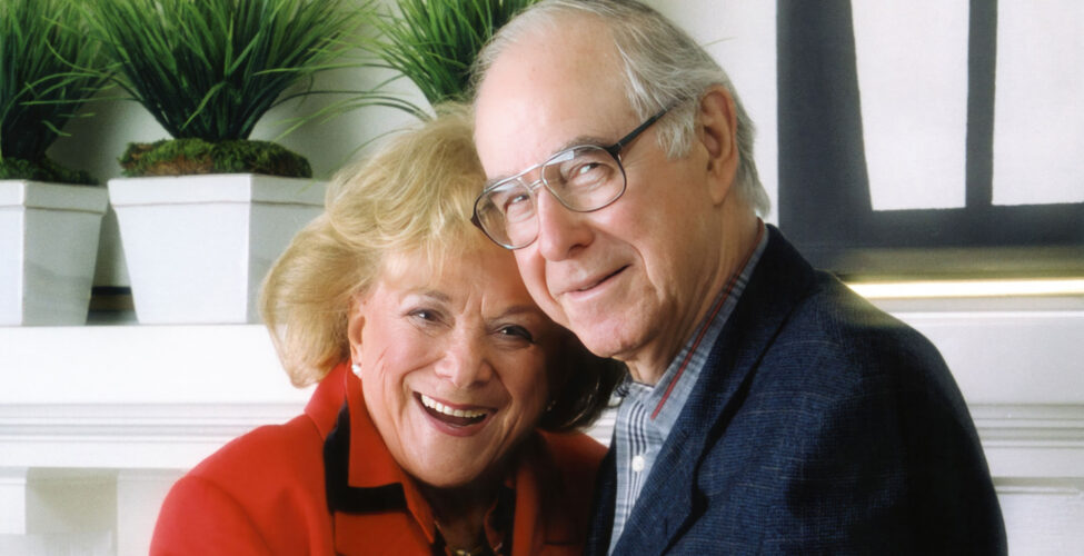 A smiling older white woman with blond hair, wearing a red jacket, standing next to a smiling older white man with eyeglasses and a blue suit coat on.
