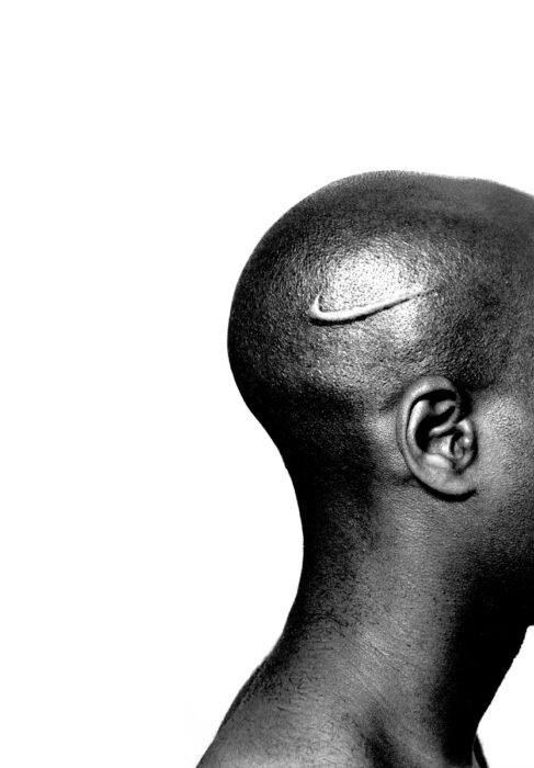 Black and white photo of a Black person's shaved head, ear, and neck with a branded Nike logo on their scalp