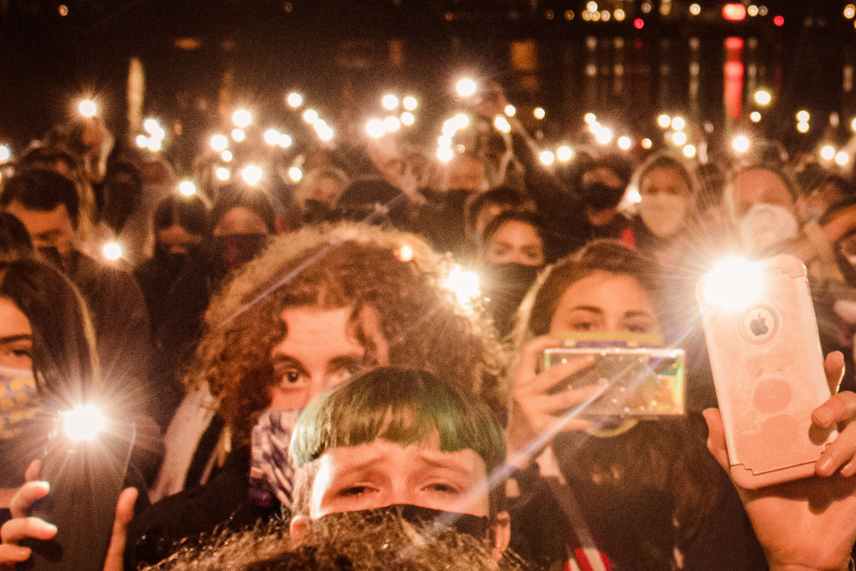 A group of people holding up their cell phones with lights on. The person in front has blue hair and tears in their eyes.