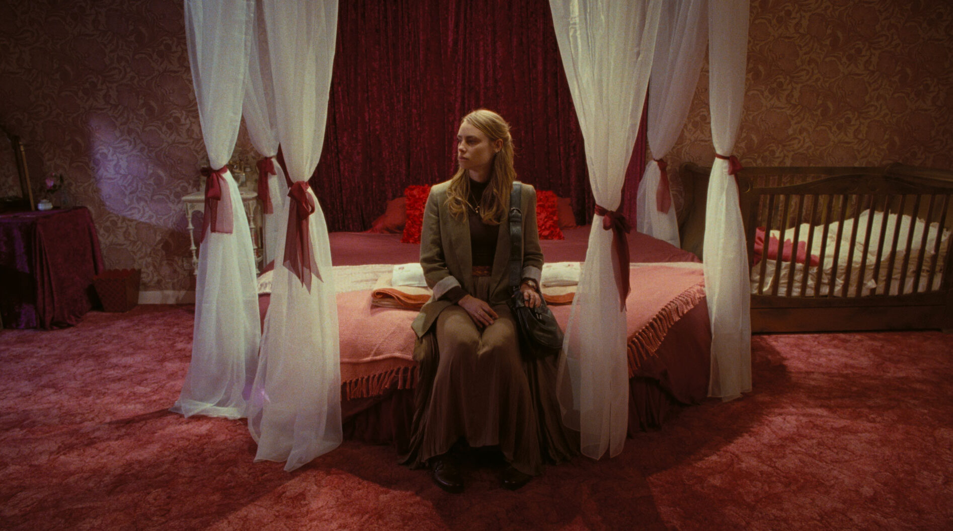 A woman sitting on a bed surrounded by curtains. There is a pink carpet and a crib next to the bed.