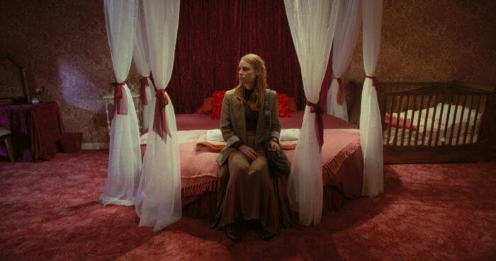 A woman sitting on a bed surrounded by curtains. There is a pink carpet and a crib next to the bed.