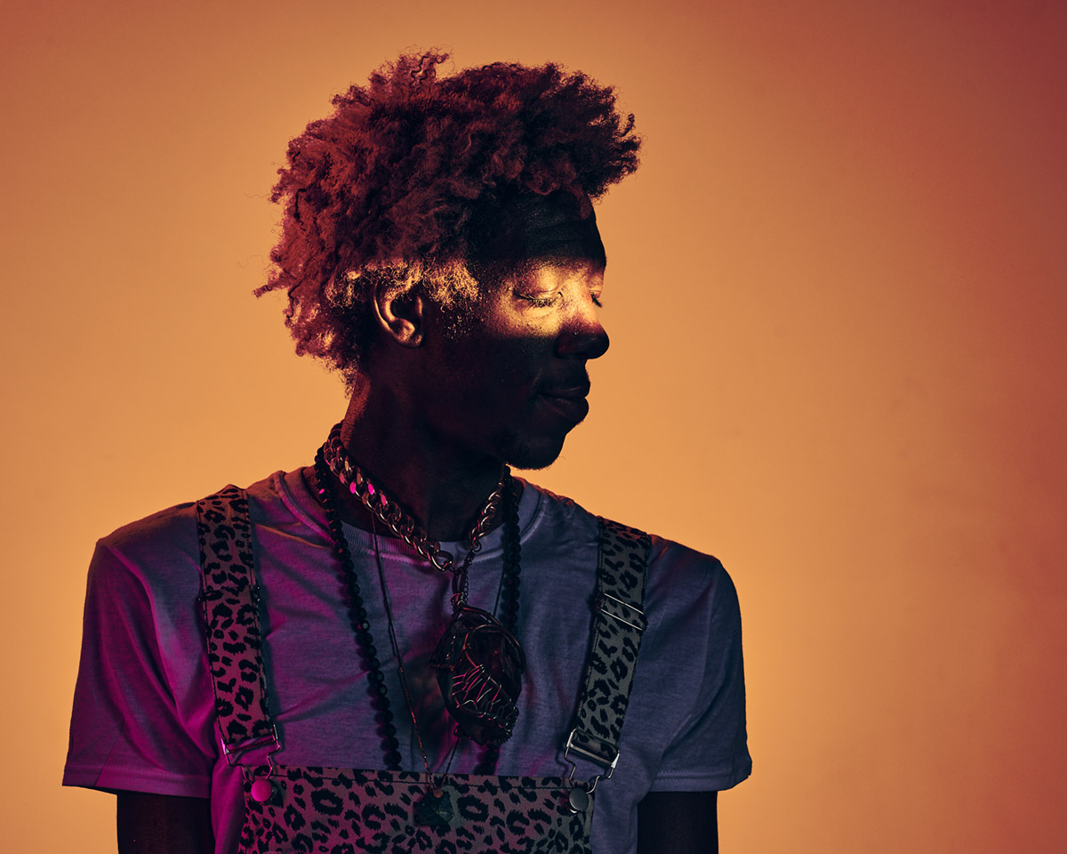 Portrait of a Black man against a orange background, looking to the side with just his closed eyes illuminated