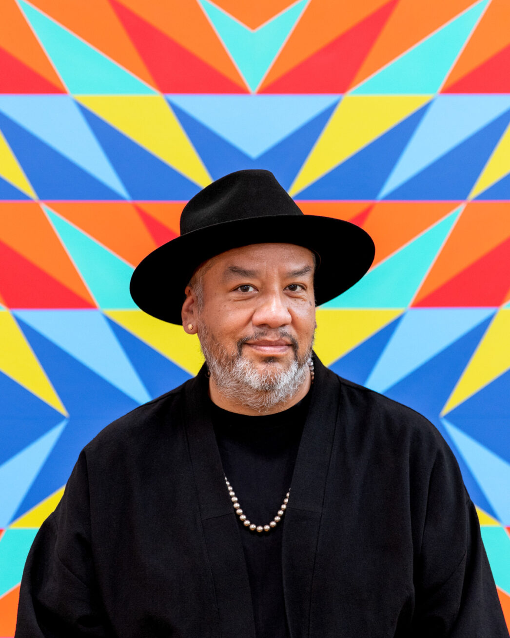 Portrait of artist Jeffrey Gibson wearing a black hat, a black shirt and jacket and a silver necklace. Standing against a colorful background.