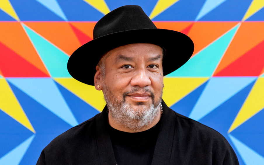 Portrait of artist Jeffrey Gibson wearing a black hat, a black shirt and jacket and a silver necklace. Standing against a colorful background.