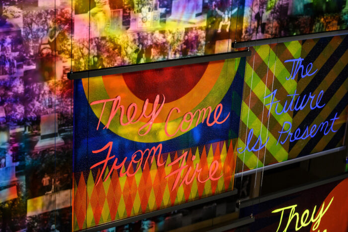 Colorful glass panel with the words "They Come From Fire" in front of a wall of black and white portraits with the reflection of the glass panel colors over them