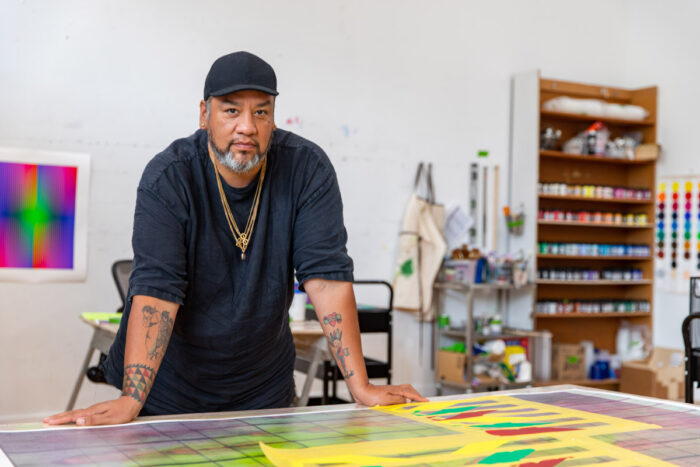 A man with a black baseball hat and a grey beard, wearing a black shirt and gold chain necklaces, standing in an art studio with both his hands resting on a table