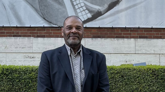 A person standing outdoors in front of a hedge.