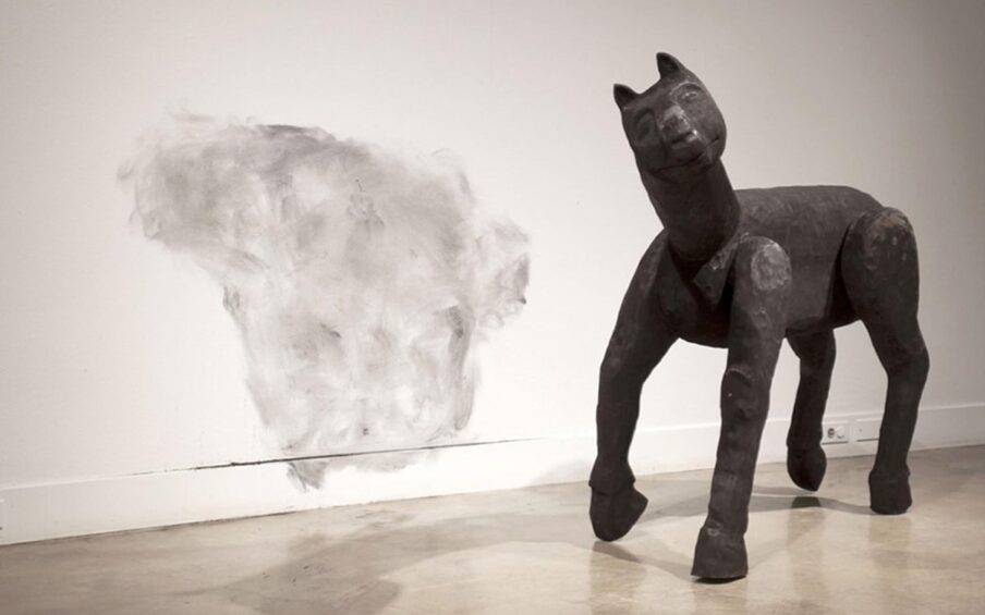 Image description: Burned Beast, Jess Perlitz, burned basswood, 48 x 30 x 54 inches. A sculpture of a four-legged, horse-like creature depicted standing with one front leg slightly raised, its head tilted to the left. The beast has a long neck, small upright ears, large deep-set eyes that appear almost closed, a short snout with a broad mouth. The creature’s legs appear to be jointed at the shoulder, and end in thick hooves. The animal is dark charcoal in color and the surface shows the uneven marks of carving. It stands directly on the floor next to a white wall that has dark smudges that roughly correlate to the beast’s silhouette.