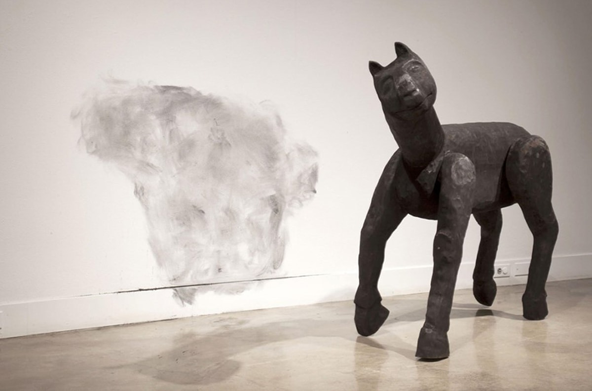 Image description: Burned Beast, Jess Perlitz, burned basswood, 48 x 30 x 54 inches. A sculpture of a four-legged, horse-like creature depicted standing with one front leg slightly raised, its head tilted to the left. The beast has a long neck, small upright ears, large deep-set eyes that appear almost closed, a short snout with a broad mouth. The creature’s legs appear to be jointed at the shoulder, and end in thick hooves. The animal is dark charcoal in color and the surface shows the uneven marks of carving. It stands directly on the floor next to a white wall that has dark smudges that roughly correlate to the beast’s silhouette.