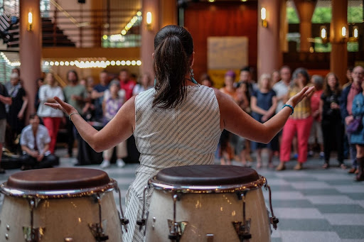 Woman with long hair in a pony tail with a bongo drum behind her, facing away from the camera toward a crowd, her arms outstretched.