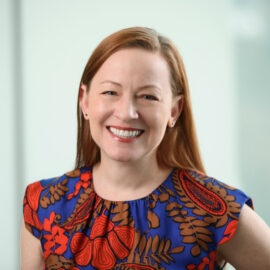 Color photograph of a woman with long, red hair smiling, wearing a bright blue, red, and brown patterned blouse